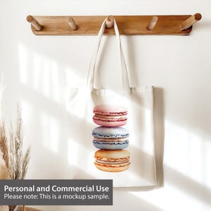 May include: A white tote bag with a design of three stacked macarons. The macarons are pink, blue, and yellow. The bag is hanging on a wooden rack with hooks.