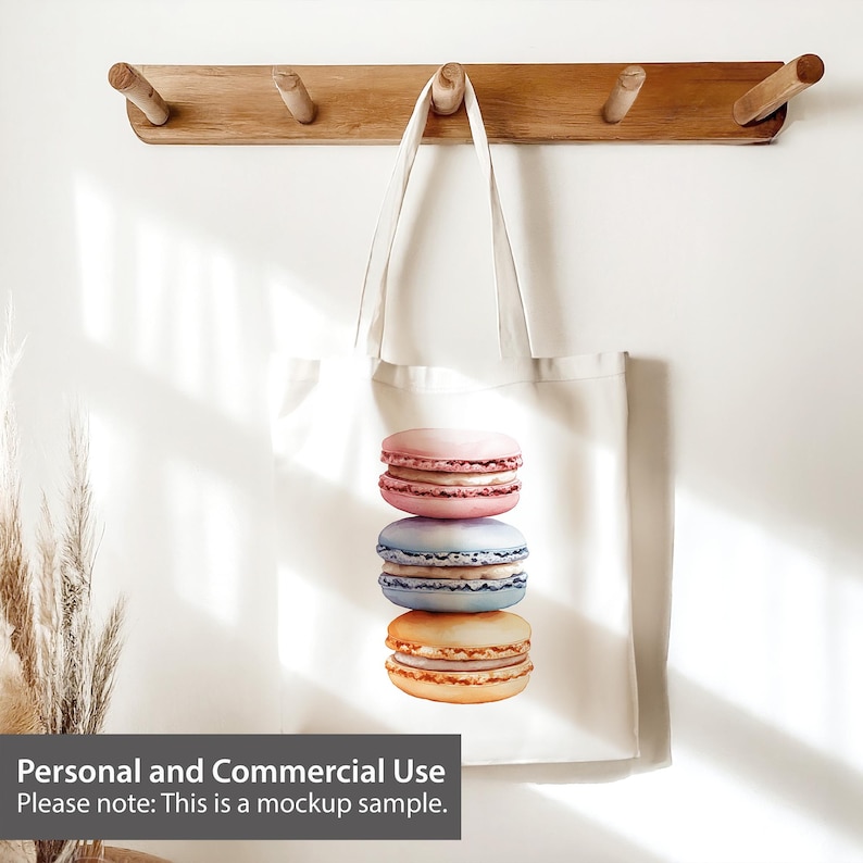 May include: A white tote bag with a design of three stacked macarons. The macarons are pink, blue, and yellow. The bag is hanging on a wooden rack with hooks.