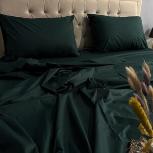 Könnte beinhalten: Dunkelgrüne Bettwäsche mit Laken und Kissenbezügen auf einem Bett mit getuftetem Kopfteil. Zwei Kissen sind sichtbar. Eine kleine schwarze Lampe und dekorative Blumen sind im Hintergrund. Die Bettwäsche ist aus einem weichen, knitterarmen Stoff.