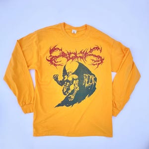 Peut inclure: T-shirt à manches longues jaune moutarde avec un motif graphique. Le motif présente un texte rouge et épineux au-dessus d'une illustration verte et noire d'une figure. Le mot "ROCK" est également visible.