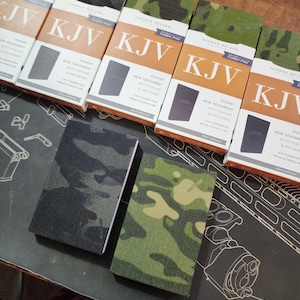 May include: Five pocket-sized New Testament KJV Bibles with Psalms and Proverbs in a camouflage print cover. The Bibles are arranged in a row from left to right, with the covers facing the viewer. The text on the covers reads "KJV Comfort Print" and "Pocket New Testament with Psalms & Proverbs".