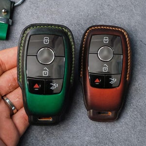 May include: Two leather key fobs in green and brown. The green fob has a black border and stitching, while the brown fob has a lighter brown border. Both have black buttons and a silver circle in the center. The key fobs are held in a hand.