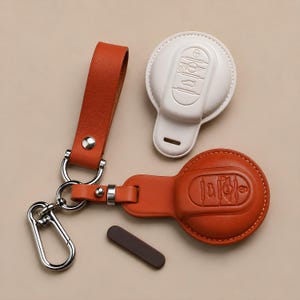 May include: An orange leather key fob cover and keychain set with a white key fob cover. The key fobs have embossed key designs. The keychain has a silver clasp and a leather strap.