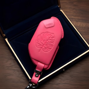 May include: A vibrant pink leather key fob case with a monstera leaf design, presented in a dark blue velvet box. The case has a matching pink strap and silver hardware. The box has a gold trim.