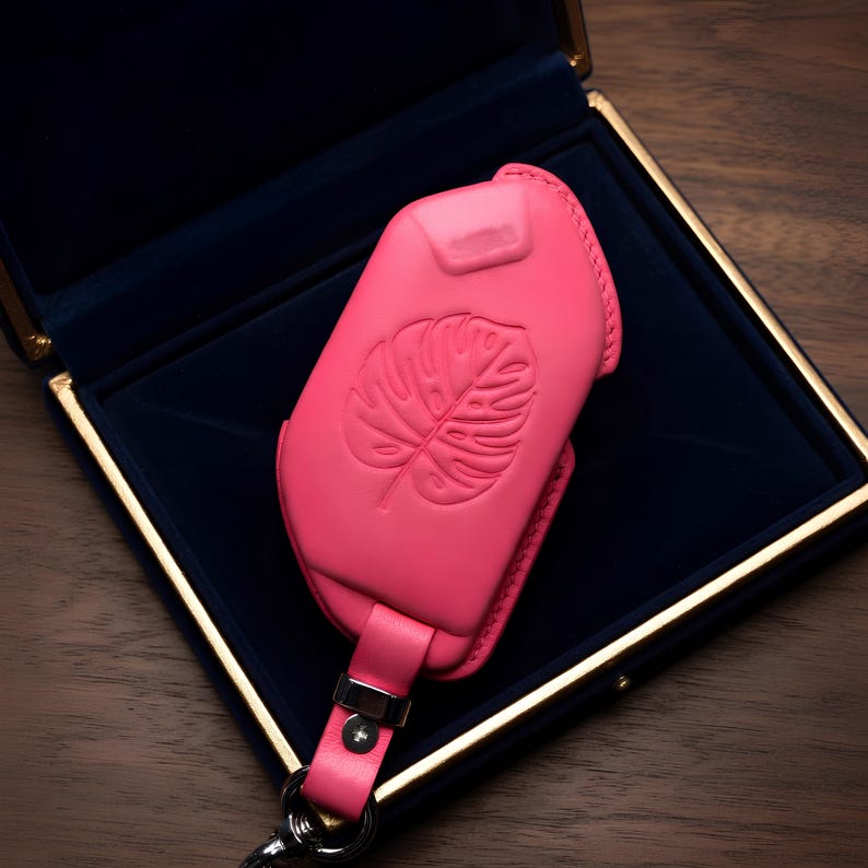 May include: A vibrant pink leather key fob case with a monstera leaf design, presented in a dark blue velvet box. The case has a matching pink strap and silver hardware. The box has a gold trim.