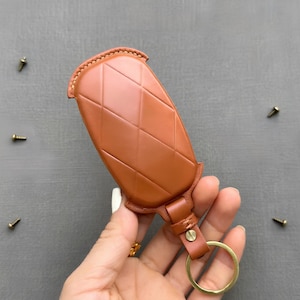 Op de afbeelding: Een bruinleren sleutelhanger met een gewatteerd patroon. De sleutelhanger heeft een goudkleurige metalen ring en een leren band. De sleutelhanger wordt in een hand gehouden tegen een grijze achtergrond.