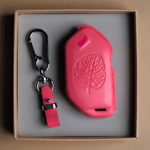 May include: A bright pink leather key fob case and key ring set. The case features a debossed monstera leaf design and a small pink accent. The key ring has a silver-tone clasp and a matching pink leather strap. The set is presented in a neutral-toned box.