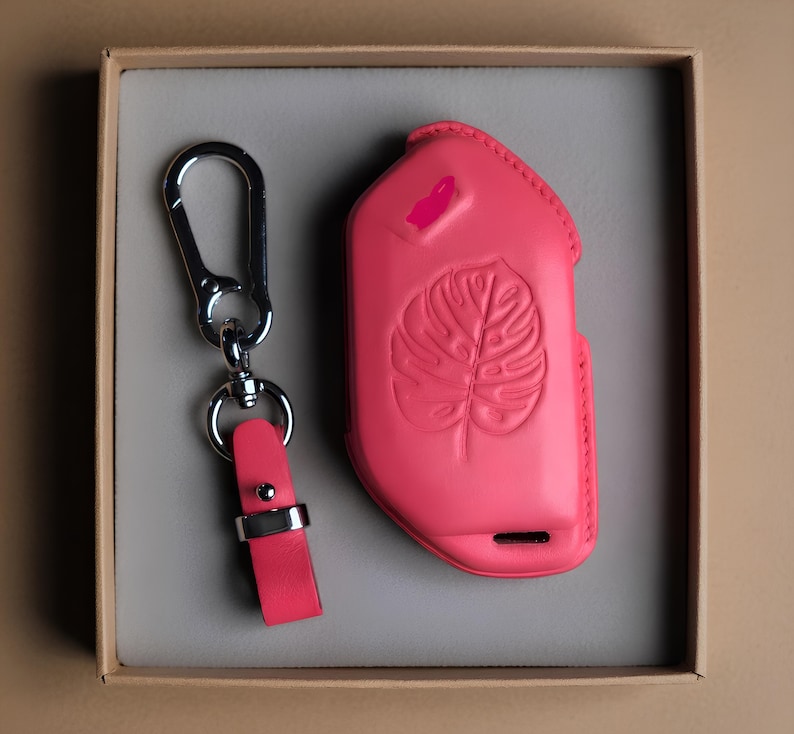 May include: A bright pink leather key fob case and key ring set. The case features a debossed monstera leaf design and a small pink accent. The key ring has a silver-tone clasp and a matching pink leather strap. The set is presented in a neutral-toned box.