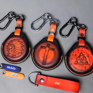 Könnte beinhalten: Handgefertigte Leder-Schlüsselanhänger mit verschiedenen geprägten Designs. Die Schlüsselanhänger zeigen einen Indianerkopfschmuck, ein Kreuz und einen keltischen Knoten. Jeder Schlüsselanhänger hat einen Metallkarabiner und einen orangefarbenen Lederriemen. Zusätzliche kleine Anhänger mit den Namen "Michelle", "XLA" und "Hall" sind enthalten.