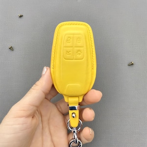 May include: A bright yellow car key fob with embossed control symbols. The fob is made of leather and has a silver key ring attached. The key fob is a vibrant yellow color and has a stitched border.