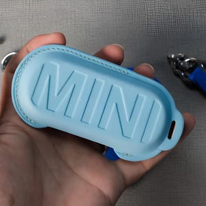May include: A light blue leather key fob case with the embossed word "MINI". The case is held in a hand, with a silver keyring and small gold-colored screws visible in the background. The case has a stitched border.