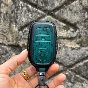 May include: A teal and black leather key fob cover with a distressed finish. The cover has embossed key symbols and a silver-tone metal clasp. The key fob is held by a hand with a gold ring.