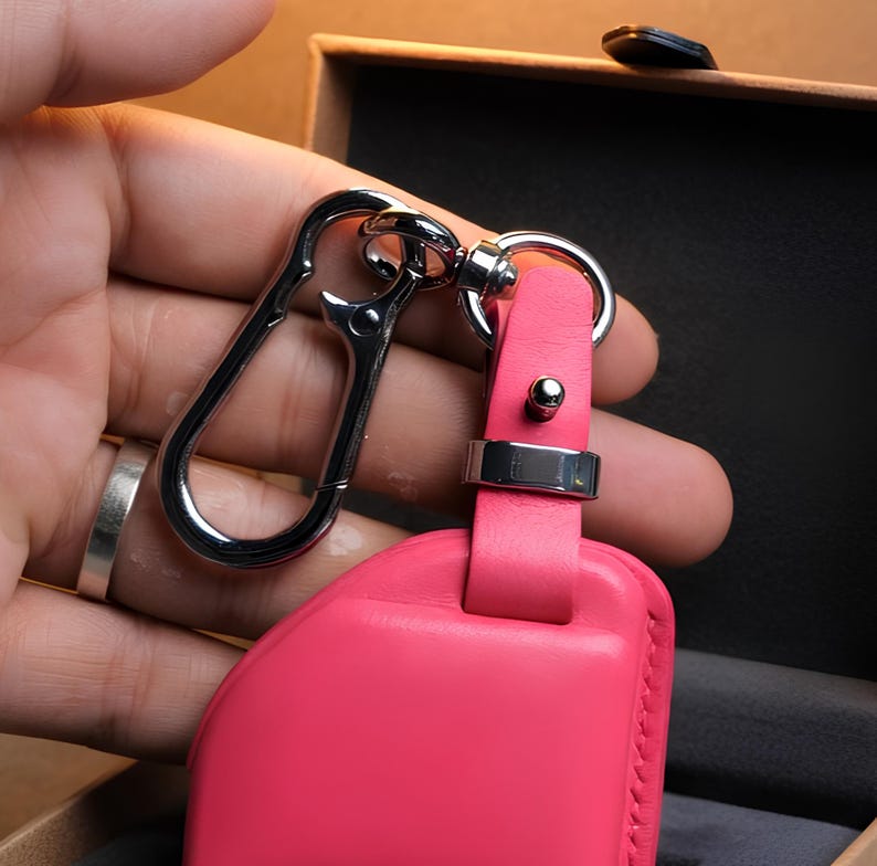 May include: A bright pink leather key fob with a silver-colored metal clasp and ring. The fob has a rectangular shape with a stitched edge. The clasp is a carabiner style, and the ring is attached to a pink leather strap.