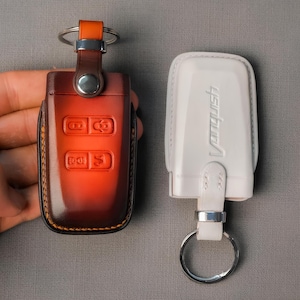 May include: Two key fobs, one in a brown leather case with orange accents and the other in a white leather case. The brown case has key function symbols. The white case has the word "Vanquish" embossed on it.