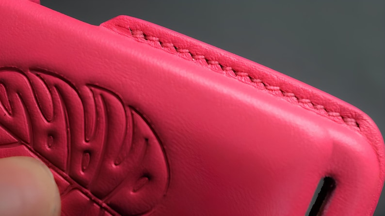 May include: Close-up of a bright pink leather phone case. The case features a stitched edge and an embossed design. The leather appears smooth and the stitching is a contrasting color. The case is a vibrant accessory.