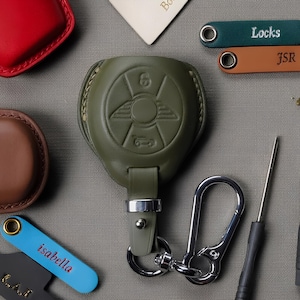 Pode incluir: Um porta-chaves de couro verde-oliva com fecho prateado, com um emblema de carro. Porta-chaves de couro adicionais em verde-azulado, castanho e azul são visíveis, com os nomes "Locks", "JSR" e "isabella" gravados. Outros acessórios e ferramentas de couro também estão presentes.