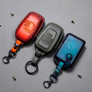 May include: Three leather key fob cases in red, olive green, and teal. The red case has an embossed eagle design, the green case reads "BAD MOTHER FUCKER", and the teal case has a button closure. Each has a metal keyring.