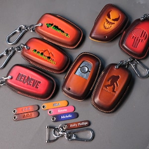 May include: A collection of leather key fob covers in various colors, including red, brown, and orange. Some covers feature embossed designs like a skull, a mountain range, and the word "BELIEVE." Also included are personalized leather tags with names.