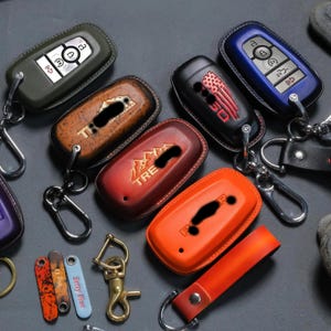 May include: A collection of leather key fob covers in various colors, including green, brown, red, black, and orange. Some covers feature embossed designs and text. Keychains and accessories are also visible.