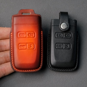 May include: Two leather key fob covers. One is a gradient of orange and brown, with visible stitching. The other is black. Both have embossed button icons and a loop for attachment.