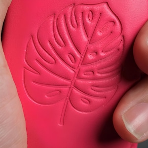 May include: Close-up of a vibrant pink object with a detailed embossed design of a Monstera leaf. The leaf's veins and shape are clearly defined. The object is held by a hand, with the focus on the textured surface and the botanical pattern.