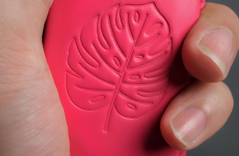 May include: Close-up of a vibrant pink object with a detailed embossed design of a Monstera leaf. The leaf's veins and shape are clearly defined. The object is held by a hand, with the focus on the textured surface and the botanical pattern.