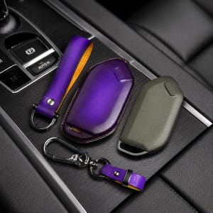 Op de afbeelding: Een paarse lederen autosleutelhouder, een grijze lederen autosleutelhouder en twee bijpassende paarse lederen sleutelhangers met zilverkleurige metalen hardware. De sleutelhoezen zijn gevormd om op voertuigsleutels te passen en hebben gladde oppervlakken.