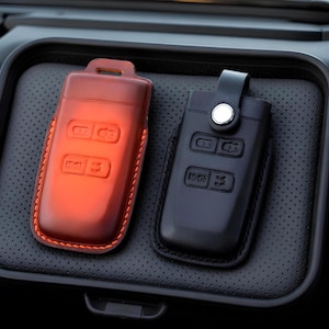 Peut inclure: Deux étuis de clés de voiture en cuir, l'un brun rougeâtre et l'autre noir, reposent sur une surface noire texturée. L'étui brun rougeâtre a des coutures orange, tandis que l'étui noir a un bouton argenté et une sangle.