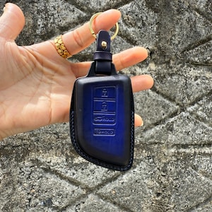 May include: A navy blue leather key fob cover with gold-colored hardware. The cover has embossed icons for lock, unlock, and hold functions. The key fob is held by a gold ring.