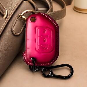 Puede incluir: Una funda de cuero magenta para la llave del coche con un mosquetón negro. La funda tiene símbolos de llave en relieve y costuras rosas. El llavero está junto a un bolso de cuero marrón.