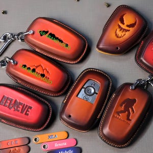 Puede incluir: Una colección de fundas de llavero de cuero en tonos marrón y rojo, con varios diseños como una calavera, una cordillera y la palabra "BELIEVE". Algunas fundas tienen llaveros adjuntos. También se ven pequeñas etiquetas con nombres.
