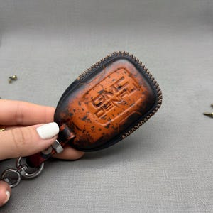 Puede incluir: Un llavero de cuero envejecido en negro y naranja, con las letras en relieve "GNE GENE". El llavero tiene un borde cosido y un accesorio de llavero de metal. El llavero está sujeto por una mano.