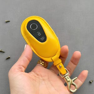 May include: A bright yellow leather key fob case with a gold-colored clasp. The case has a matching yellow leather strap and a key ring. The key fob has a black panel with white icons.