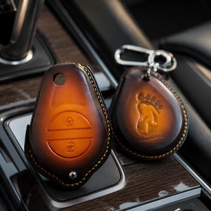 May include: Two leather key fobs with a gradient brown finish. One is a key case with lock and unlock symbols. The other is a teardrop shape with a footprint design and a silver keyring.