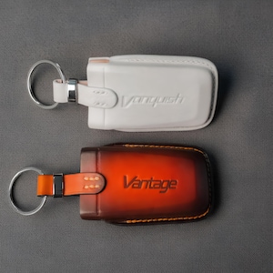 May include: Two leather keychains. The top one is white with the word "Vanquish" embossed. The bottom keychain is brown and orange with the word "Vantage" embossed. Both have a metal ring and strap.