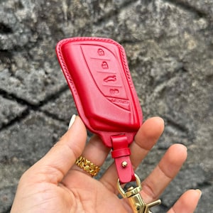 Puede incluir: Una funda roja de cuero para la llave del coche con botones en relieve y el logotipo de Lexus. El llavero tiene una correa de cuero y un cierre dorado. El fondo es una superficie gris texturizada.