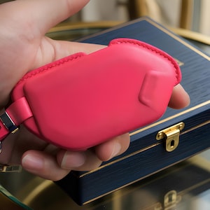 May include: A vibrant pink leather key fob case with a silver-tone clasp and key ring. The case is held in a hand, with a dark blue wooden box with gold trim and a clasp in the background.
