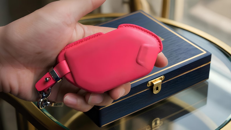 May include: A vibrant pink leather key fob case with a silver-tone clasp and key ring. The case is held in a hand, with a dark blue wooden box with gold trim and a clasp in the background.
