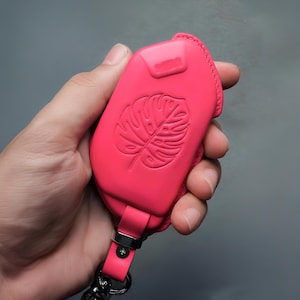 Peut inclure: Un étui de porte-clés en cuir rose vif avec un motif de feuille de monstera en relief. L'étui a une sangle assortie et une quincaillerie argentée. L'étui de porte-clés est tenu dans une main sur un fond gris.