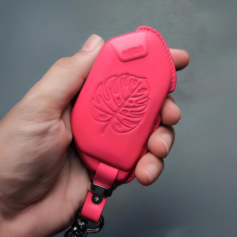 May include: A bright pink leather key fob case with a debossed monstera leaf design. The case has a matching strap and silver-tone hardware. The key fob case is held in a hand against a gray background.