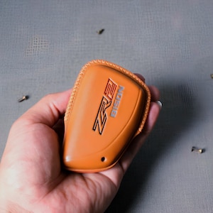 May include: A tan leather key fob cover with the text "ZR2 BISON" in black and blue. The cover is rectangular with rounded edges and a stitched border. The key fob cover is held in a hand.