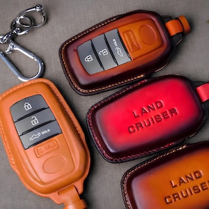 Può includere: Primo piano di diverse custodie per portachiavi in pelle nei toni del marrone e dell'arancione. Una custodia è rosso vivo con la scritta "LAND CRUISER" in rilievo. Sono visibili anche portachiavi con finiture argentate.