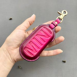 May include: A magenta leather key fob with the word "MINI" embossed on the front. The fob has a gold-coloured clasp and a matching leather strap. The key fob is designed to hold a car key.
