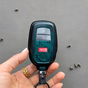 May include: A dark teal, distressed leather key fob cover featuring a black border and stitching. The cover has cutouts for three buttons: a lock icon, an unlock icon, and a red button labeled HOLD. A black metal ring is attached at the base.