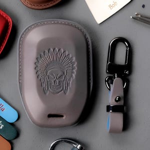 Puede incluir: Una funda para llavero de cuero gris con un diseño de calavera y tocado. Un mosquetón de metal negro está unido a una correa de cuero gris. También se ven otros artículos de cuero y etiquetas personalizadas.