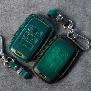May include: Two teal leather key fob covers with silver keychains. The covers have embossed button icons and a dark border. One keychain has a carabiner clip, the other a split ring. The key fobs are on a grey surface.