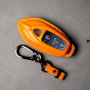 May include: An orange leather key fob cover with a silver carabiner and a small orange leather strap. The key fob has three buttons with white icons. The key fob cover is on a gray surface.