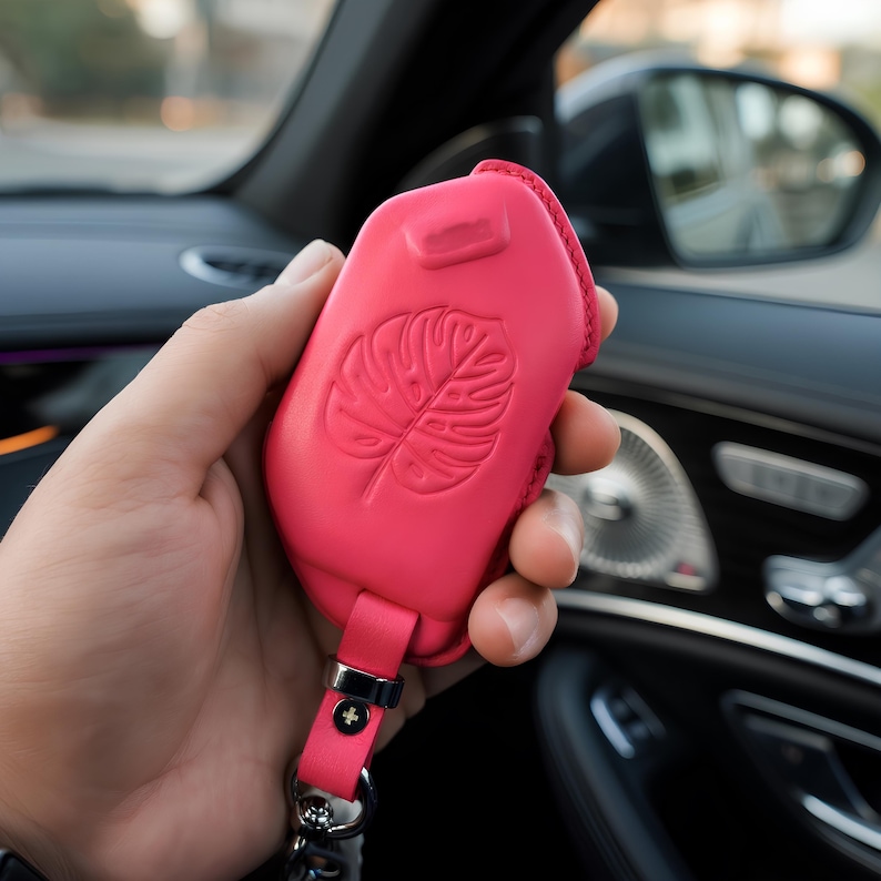May include: A vibrant pink car key fob cover with a leaf design. The leather case has a matching strap and silver-tone hardware. The key fob is held in a car interior.
