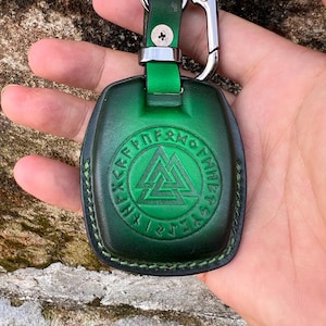 Puede incluir: Llavero de cuero verde con cierre plateado. El llavero presenta un diseño circular en relieve con runas y un símbolo triangular central. El cuero está cosido alrededor de los bordes y tiene una correa de cuero verde.