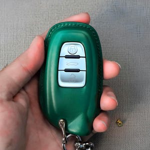 May include: A green leather car key fob cover with silver buttons and a key ring. The fob has a lock, car, and trunk button. The cover is held in a hand.